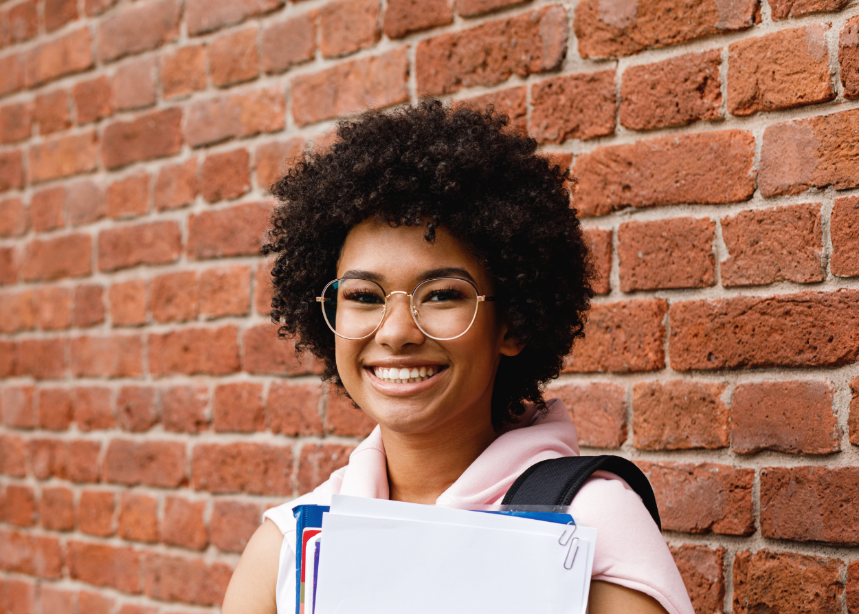 smiling student carrying textbooks standing in front of brick wall